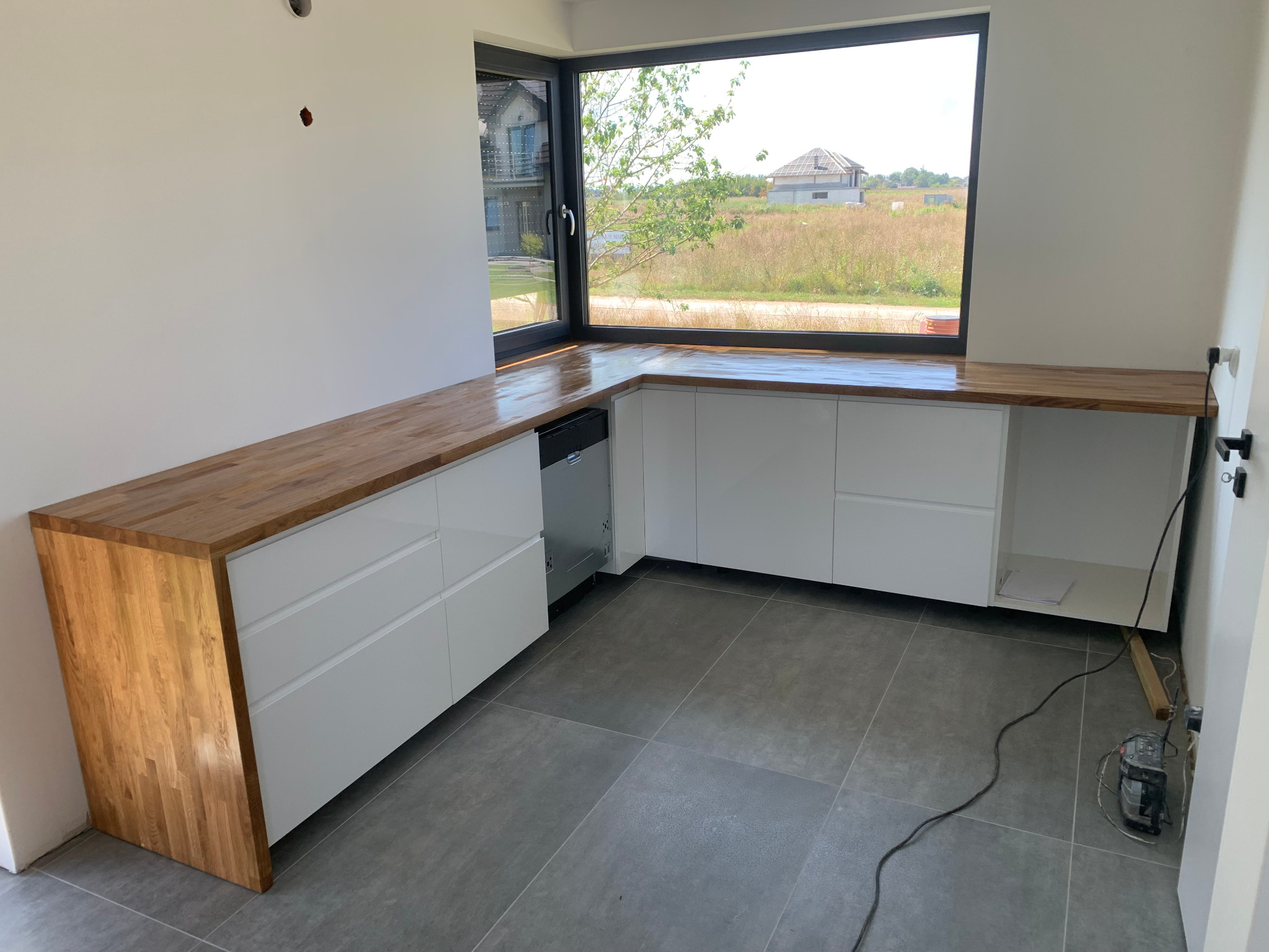 Nowoczesna kuchnia w trakcie montażu: białe szafki z drewnianym blatem, widok na krajobraz za oknem, szare płytki podłogowe, narzędzia na podłodze.