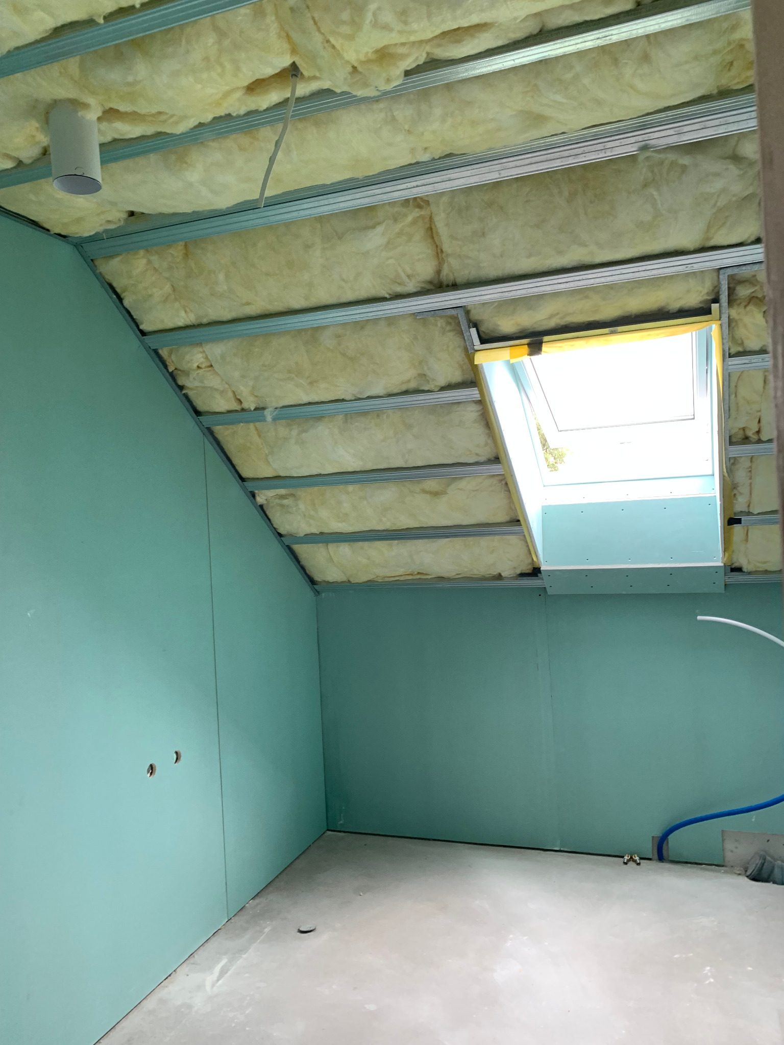 Poddasze w trakcie adaptacji: ocieplony sufit z wełny mineralnej, metalowa konstrukcja nośna, ściany obłożone płytami gipsowo-kartonowymi w kolorze miętowym, okno dachowe z żółtą ramą, widoczne...