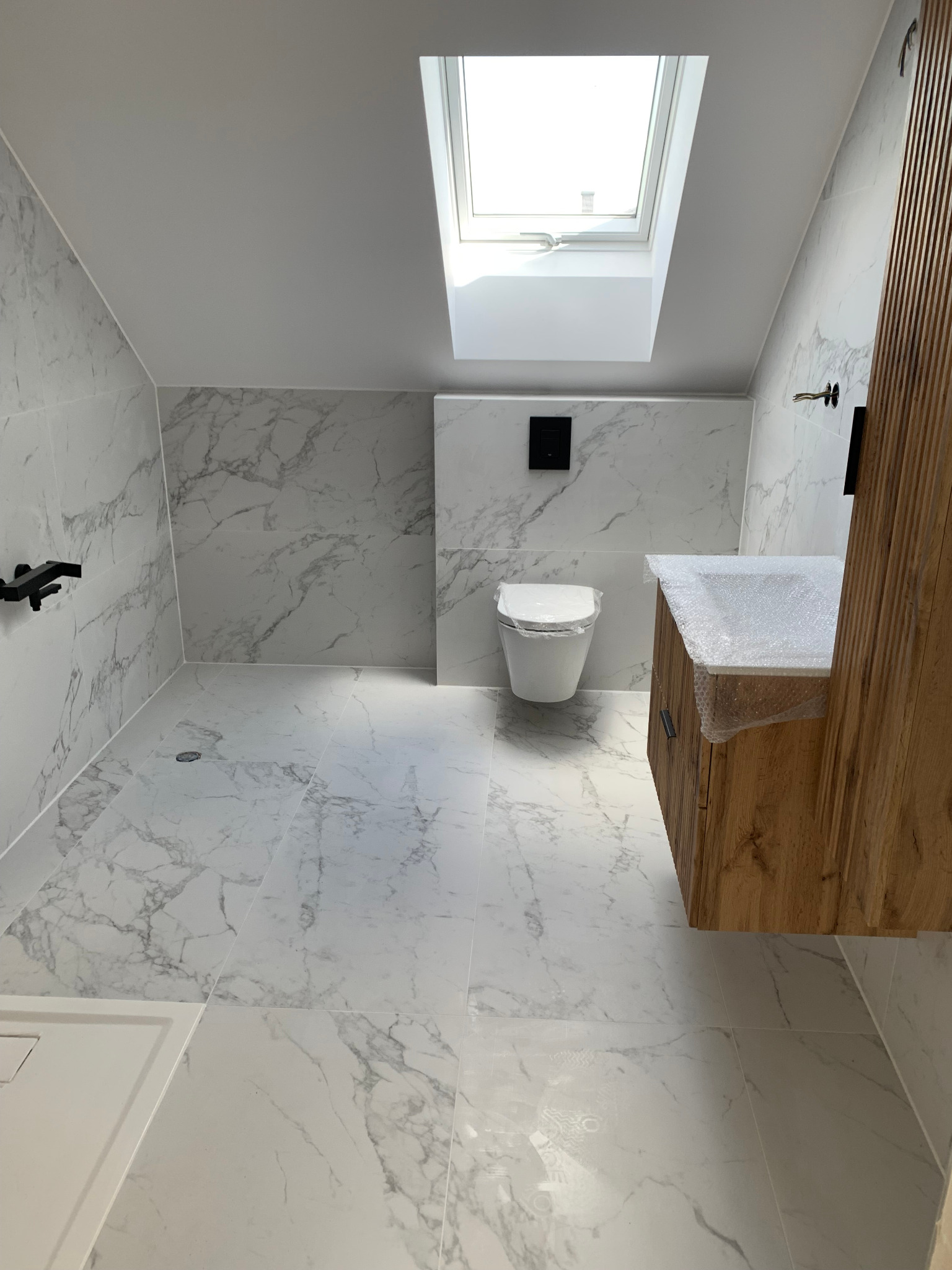 Nowoczesna łazienka na poddaszu z marmurowymi płytkami na ścianach i podłodze, drewniana szafka pod umywalką częściowo zabezpieczona folią bąbelkową, wisząca toaleta i okno dachowe wpuszczające...