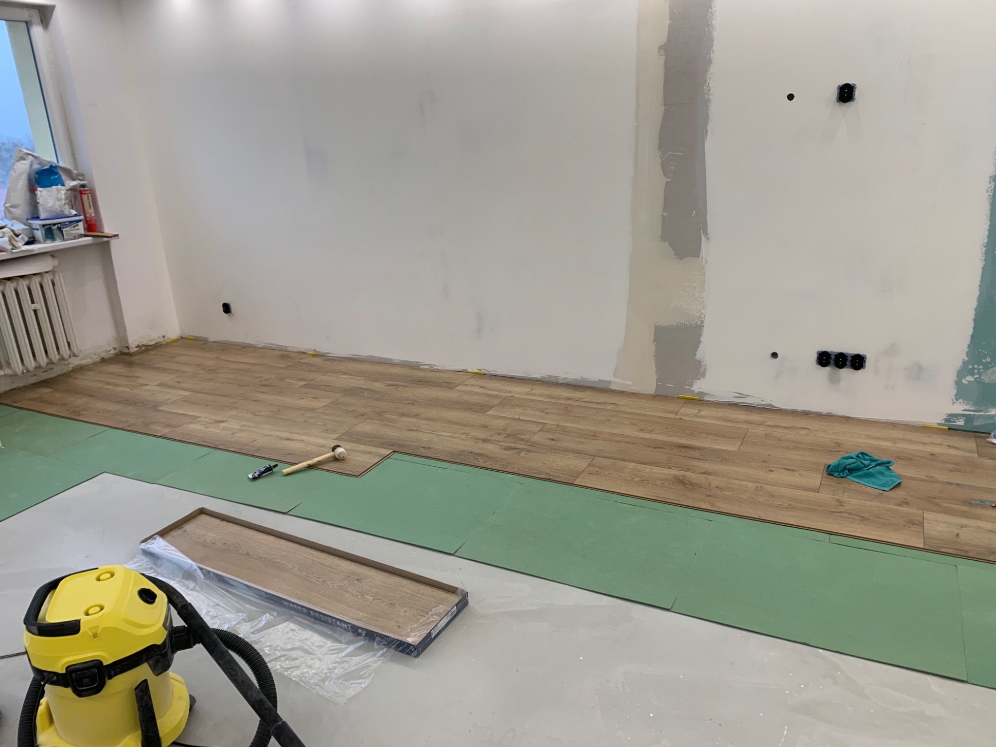 Układanie paneli podłogowych w pokoju, widoczna podkładka izolacyjna, paczka paneli, gumowy młotek, żółty odkurzacz przemysłowy i fragment ściany przygotowanej do malowania.