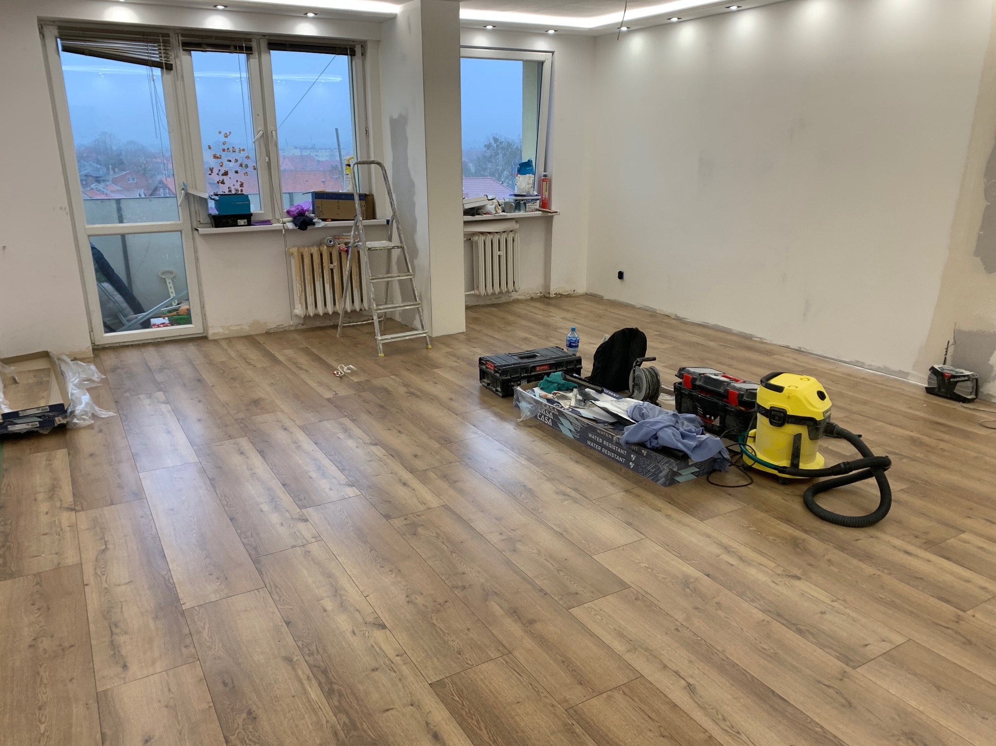 Pokój w trakcie prac wykończeniowych: świeżo położona podłoga laminowana, drabina oparta o ścianę, narzędzia i odkurzacz przemysłowy w tle, ściany przygotowane do malowania.