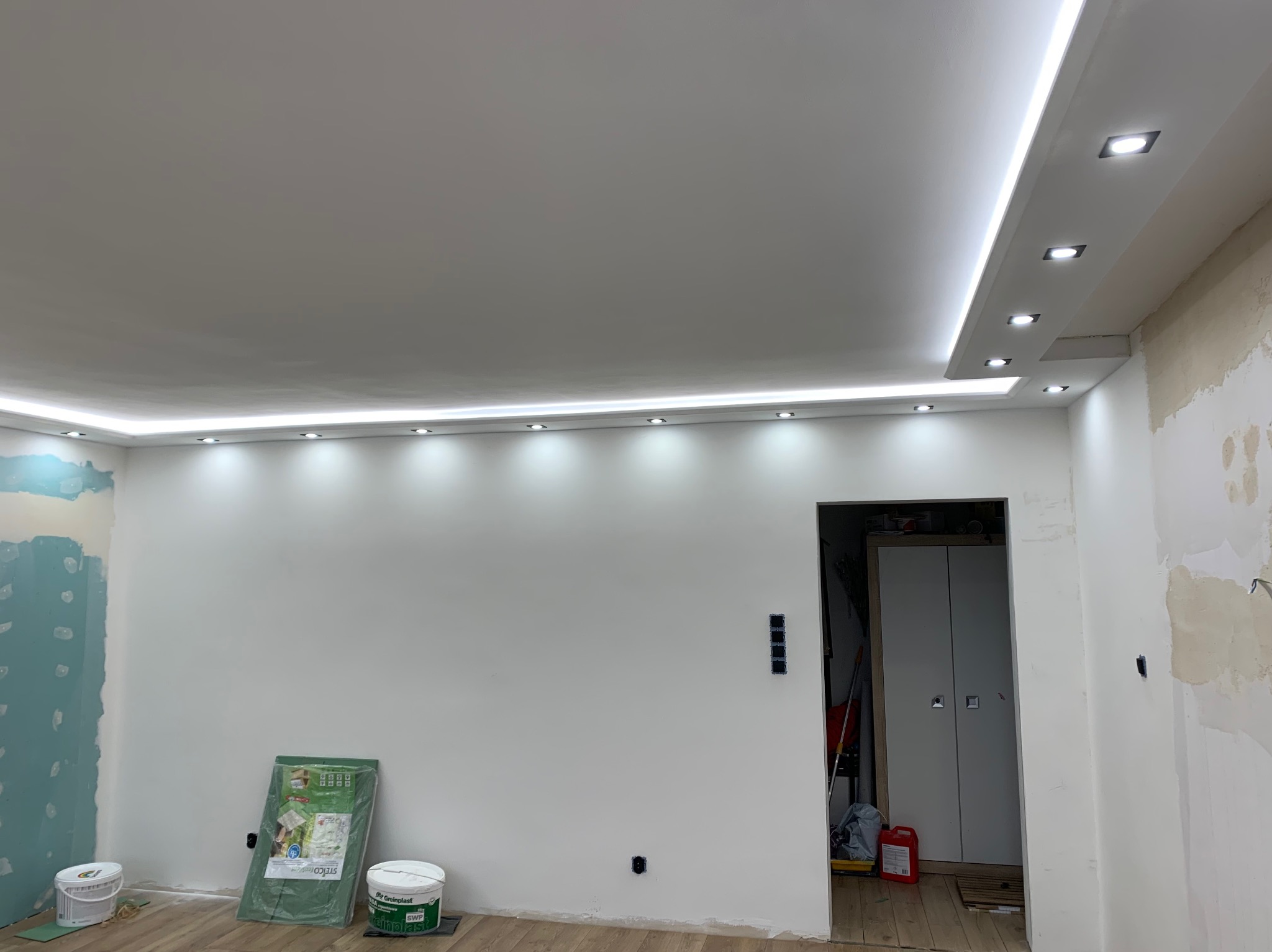 Wnętrze w trakcie prac wykończeniowych, widoczny sufit podwieszany z oświetleniem LED, ściany częściowo przygotowane do malowania, puszki elektryczne, wiadro z farbą i folia ochronna na podłodze.
