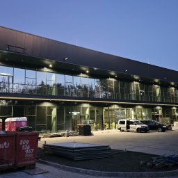 AVISTAL SPÓŁKA Z OGRANICZONĄ ODPOWIEDZIALNOŚCIĄ - Dwukondygnacyjny budynek z płyt warstwowych, widoczny kontener na gruz i pracownicy w odblaskowych kamizelkach. Nowoczesna architektura, wieczorne oświetlenie.