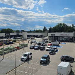 AVISTAL SPÓŁKA Z OGRANICZONĄ ODPOWIEDZIALNOŚCIĄ - Panorama centrum handlowego w budowie z widocznymi sklepami, parkingiem i materiałami budowlanymi na zewnątrz, przy słonecznej pogodzie.