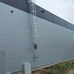 AVISTAL SPÓŁKA Z OGRANICZONĄ ODPOWIEDZIALNOŚCIĄ - Szara hala z płyt warstwowych z drabiną ewakuacyjną, widok z dołu. Obok fragment konstrukcji z okładziną 3XL. Widoczne elementy instalacji elektrycznej.