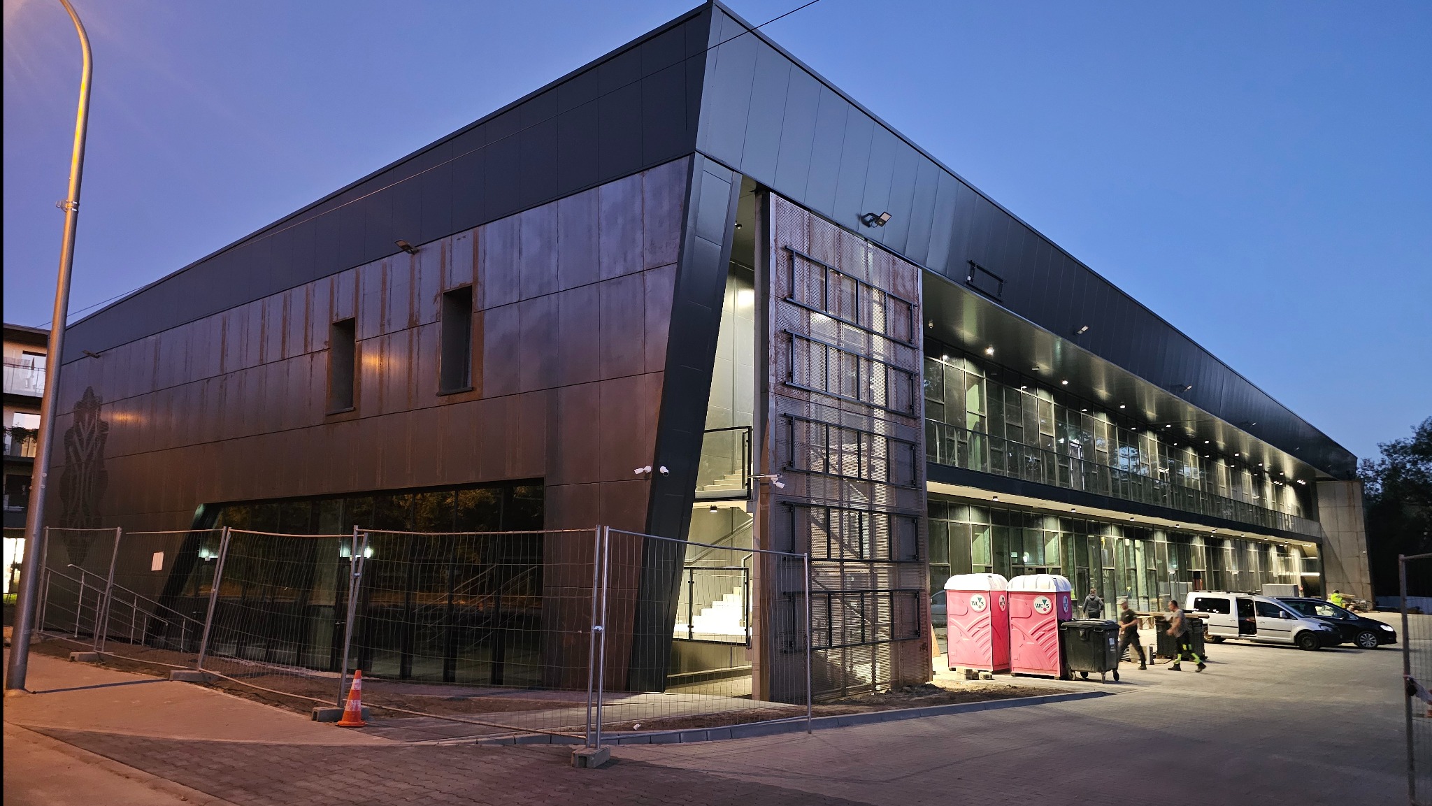 Nowoczesny budynek biurowy z płyt warstwowych w szaro-brązowej kolorystyce, oświetlony wieczornym światłem, z widocznymi detalami wykończenia i elementami infrastruktury.