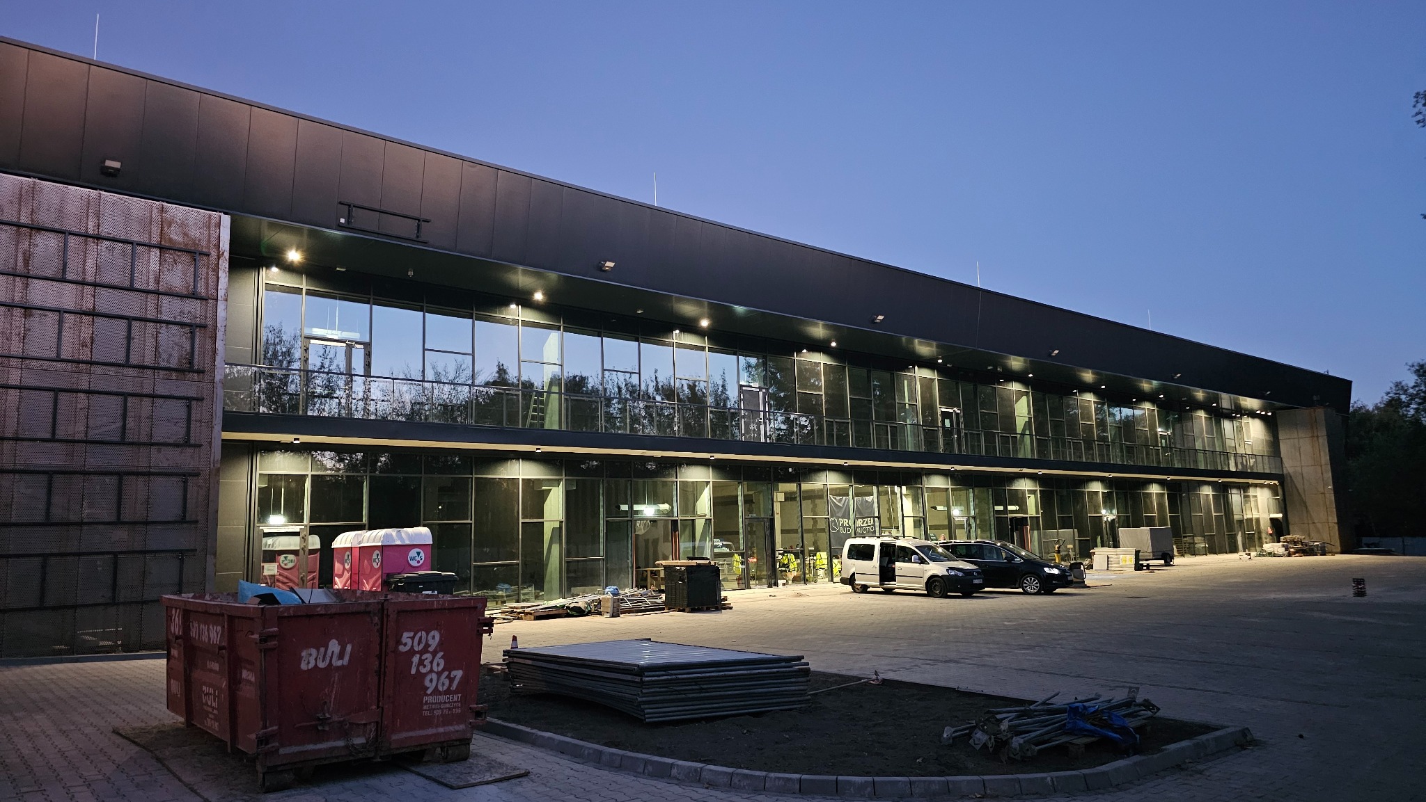 Dwukondygnacyjny budynek z płyt warstwowych, widoczny kontener na gruz i pracownicy w odblaskowych kamizelkach. Nowoczesna architektura, wieczorne oświetlenie.