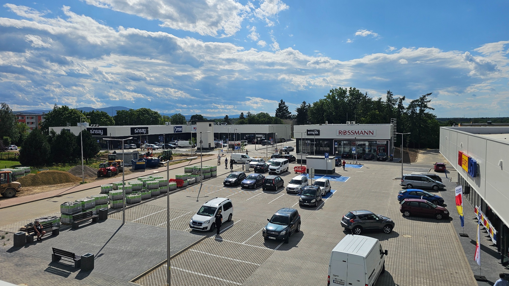 Panorama centrum handlowego w budowie z widocznymi sklepami, parkingiem i materiałami budowlanymi na zewnątrz, przy słonecznej pogodzie.