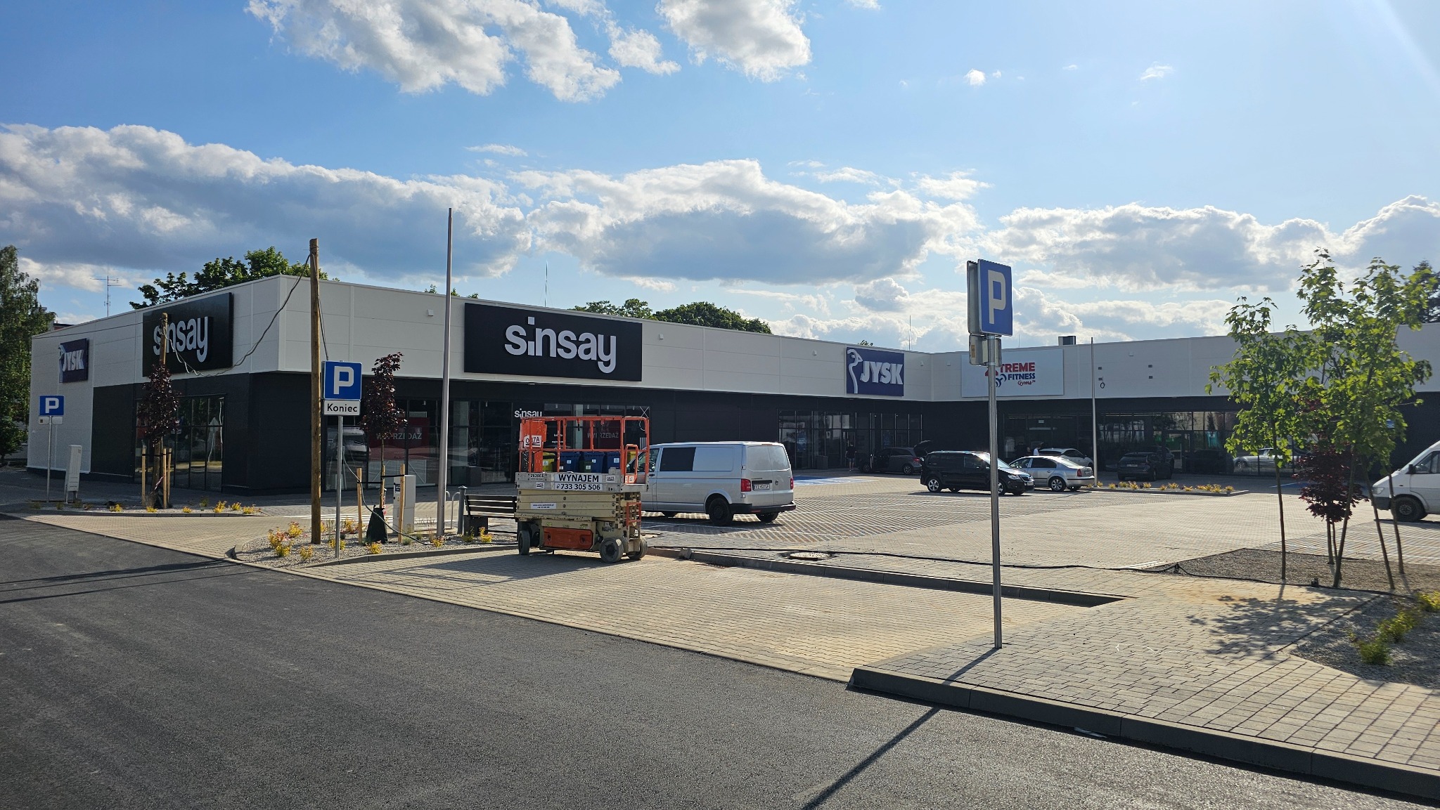 Budynek handlowy z płyt warstwowych, widoczny podnośnik koszowy. Fasada z logo sklepów JYSK i Sinsay. Parking przed budynkiem, błękitne niebo z chmurami.