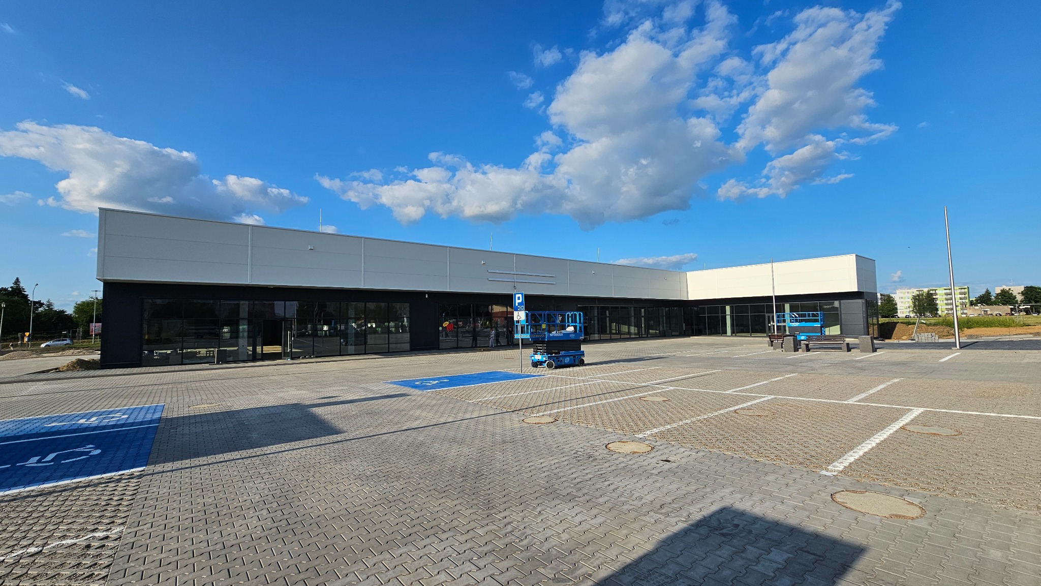 Nowoczesny budynek z płyt warstwowych, widok z zewnątrz. Na pierwszym planie podnośnik koszowy. Błękitne niebo z chmurami w tle.