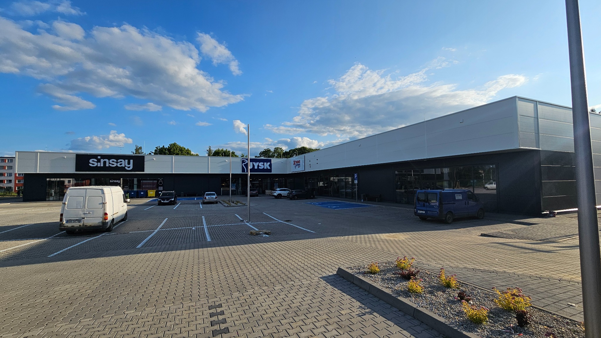 Budynek handlowy z elewacją z płyt warstwowych, widoczne sklepy JYSK i Sinsay, parking z zaparkowanymi samochodami i dostawczymi busami, błękitne niebo.