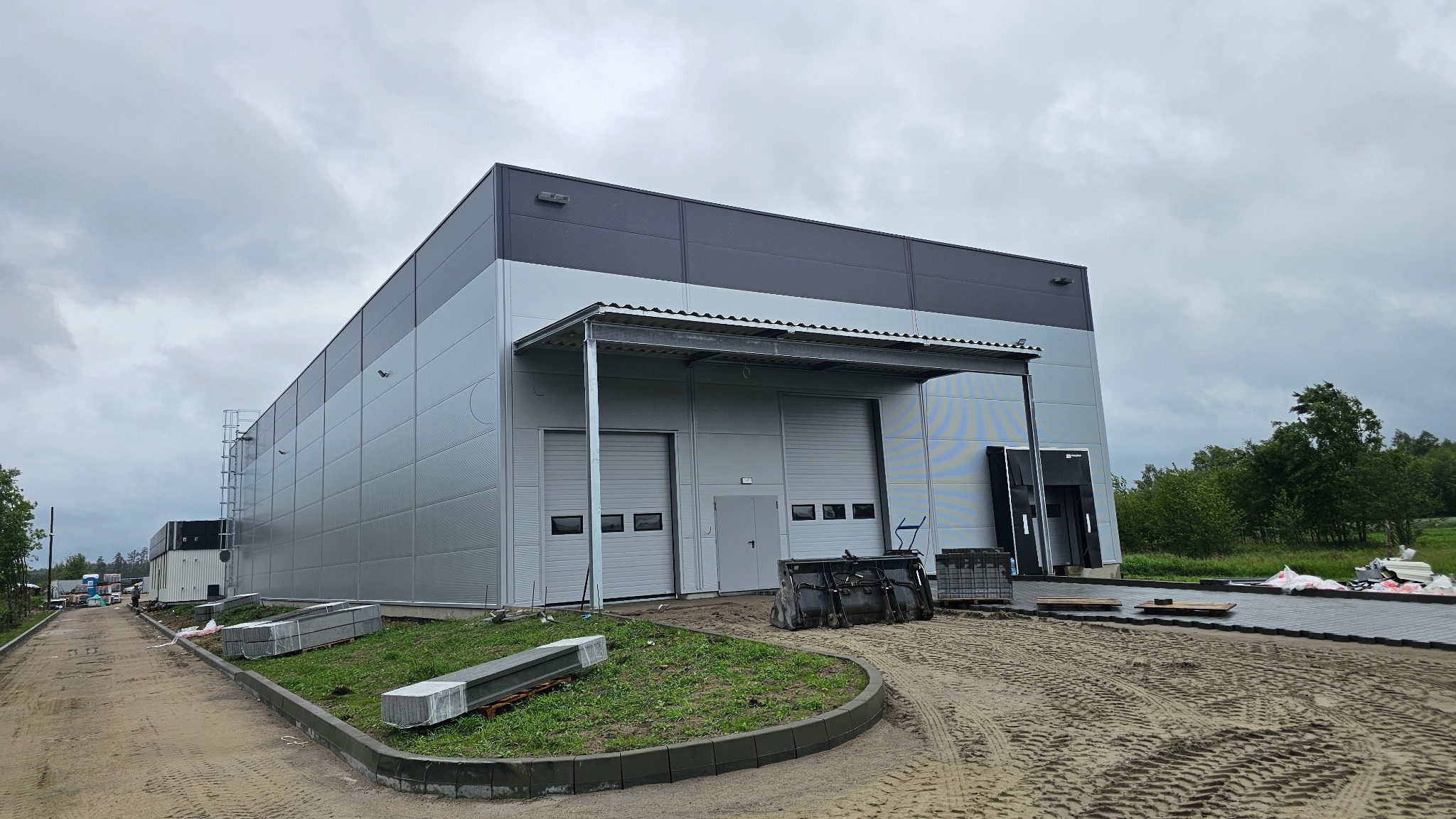 Hala z płyt warstwowych w odcieniach szarości, z widocznymi dwoma bramami garażowymi i fragmentem terenu budowy. Pochmurne niebo w tle.