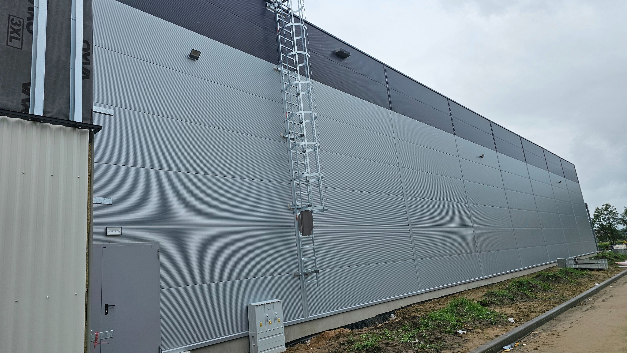 Szara hala z płyt warstwowych z drabiną ewakuacyjną, widok z dołu. Obok fragment konstrukcji z okładziną 3XL. Widoczne elementy instalacji elektrycznej.
