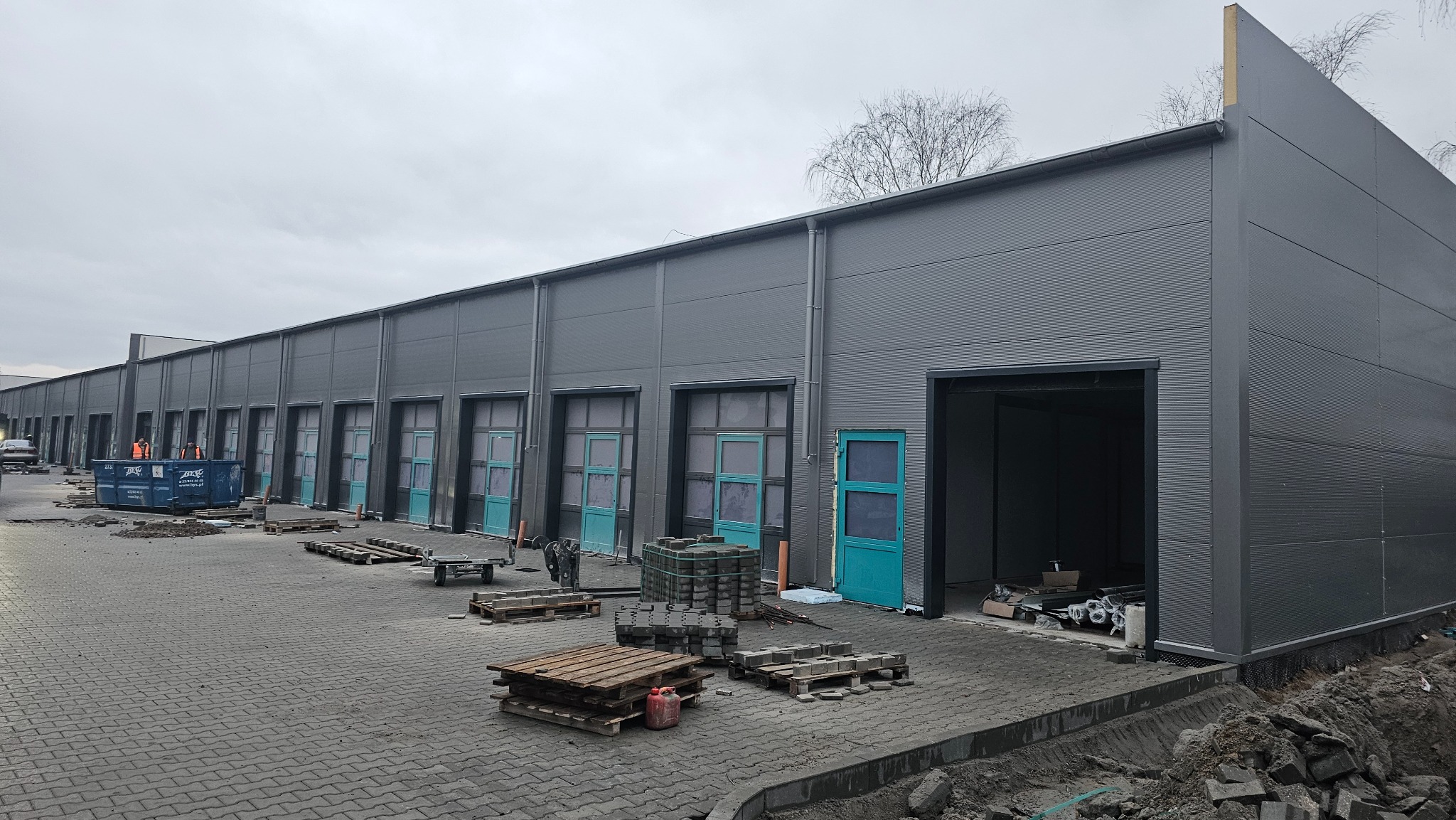 Hala z płyt warstwowych w trakcie budowy, widoczne turkusowe drzwi i okna, szara elewacja, teren budowy z kostki brukowej, palety i kontener na odpady.