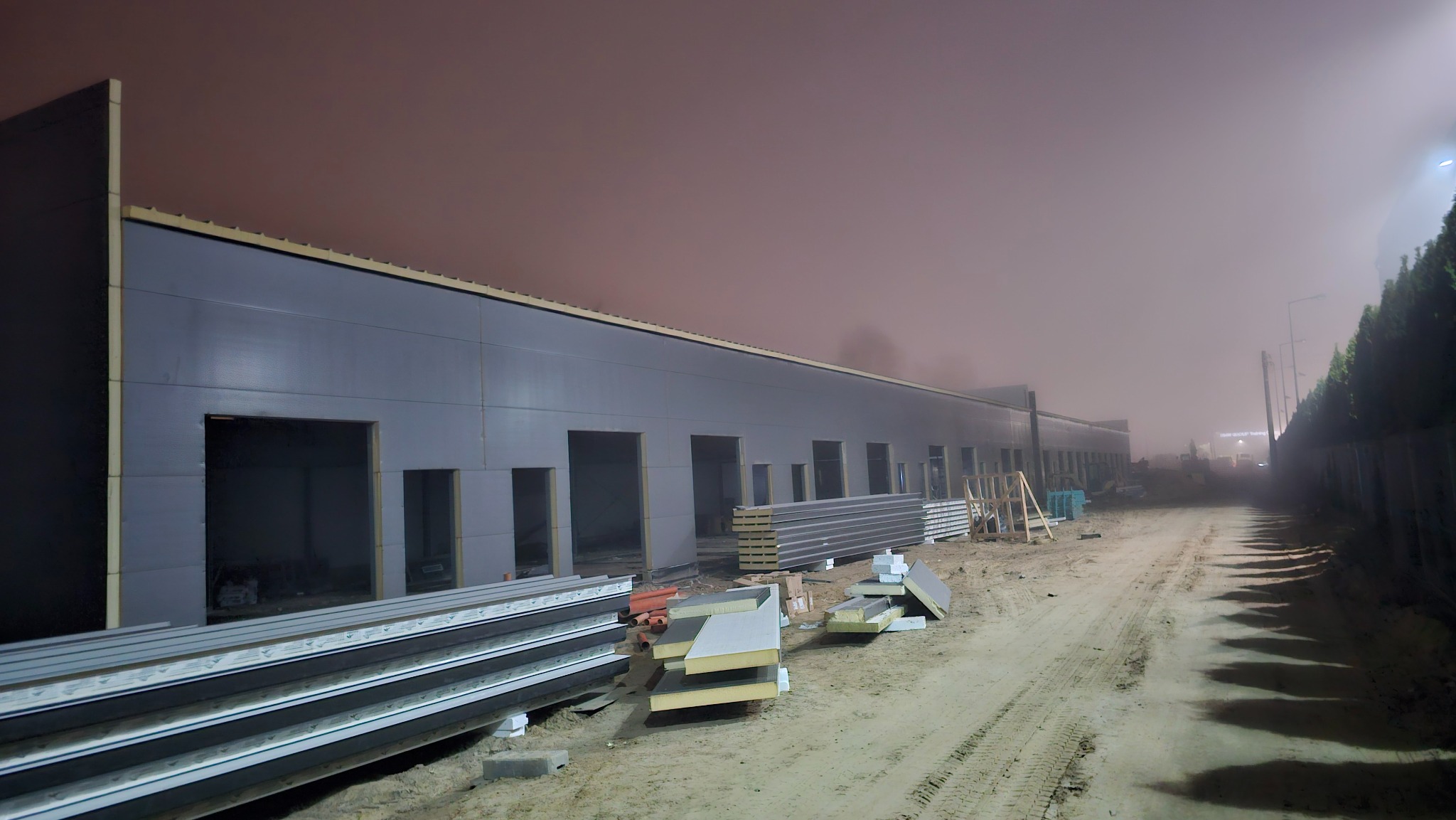 Nocny montaż płyt warstwowych na hali przemysłowej w trakcie budowy, widoczne stosy płyt i otwory okienne w szarej elewacji, oświetlone lampami ulicznymi.
