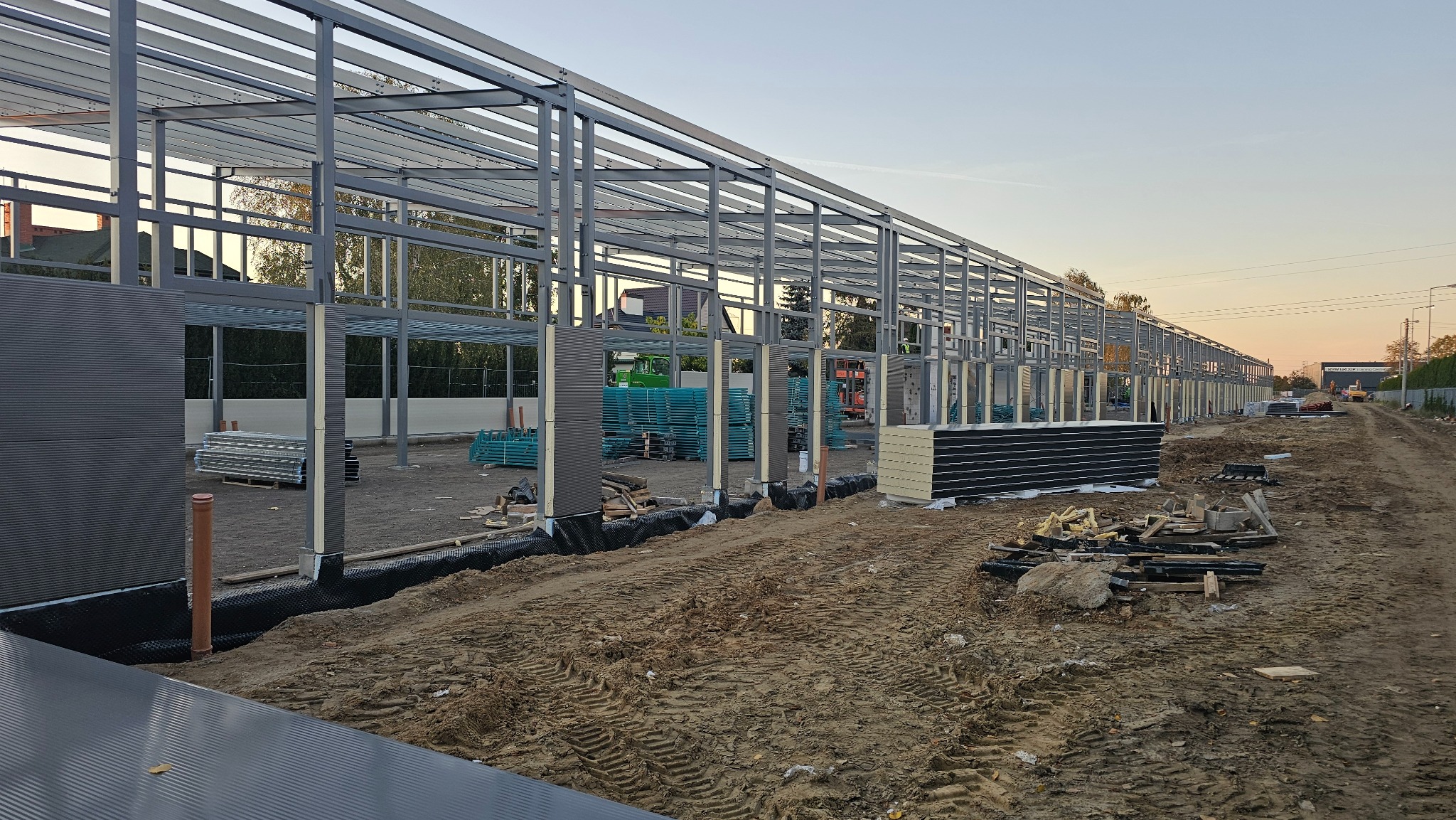 Konstrukcja stalowa hali w trakcie montażu płyt warstwowych, widoczne elementy konstrukcyjne i przygotowane płyty. W tle sprzęt budowlany i zachodzące słońce.