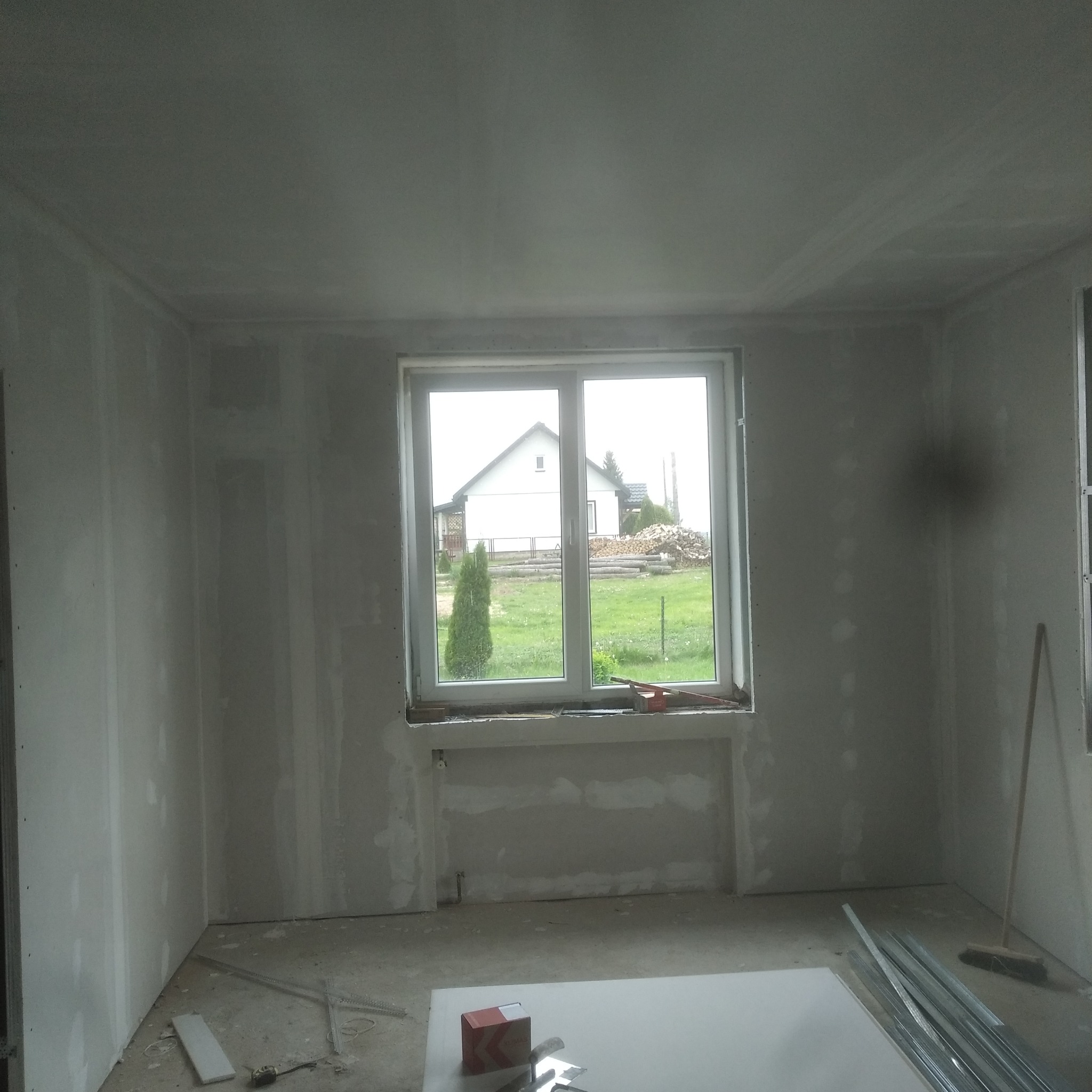 Wnętrze w trakcie prac wykończeniowych: ściany pokryte płytami gipsowo-kartonowymi, widoczne profile metalowe, narzędzia i materiały budowlane na podłodze, okno z widokiem na ogród.