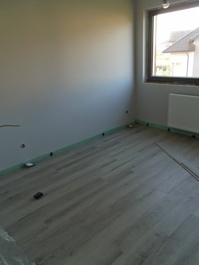 Pokój z ułożonymi panelami podłogowymi w odcieniach szarości, zabezpieczony folią przy ścianach, widoczne otwory na gniazdka elektryczne i grzejnik pod oknem.