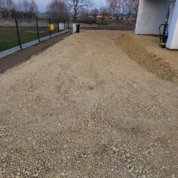 Teren przygotowany pod przyszłą nawierzchnię z kostki brukowej, widoczna warstwa podsypki z kruszywa, obrzeża betonowe i częściowo ogrodzenie panelowe, w tle budynek mieszkalny w stanie surowym...