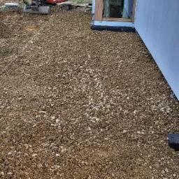 Przygotowanie terenu pod budowę: mini koparka Kubota i warstwa tłucznia przy ścianie domu z dużym oknem.