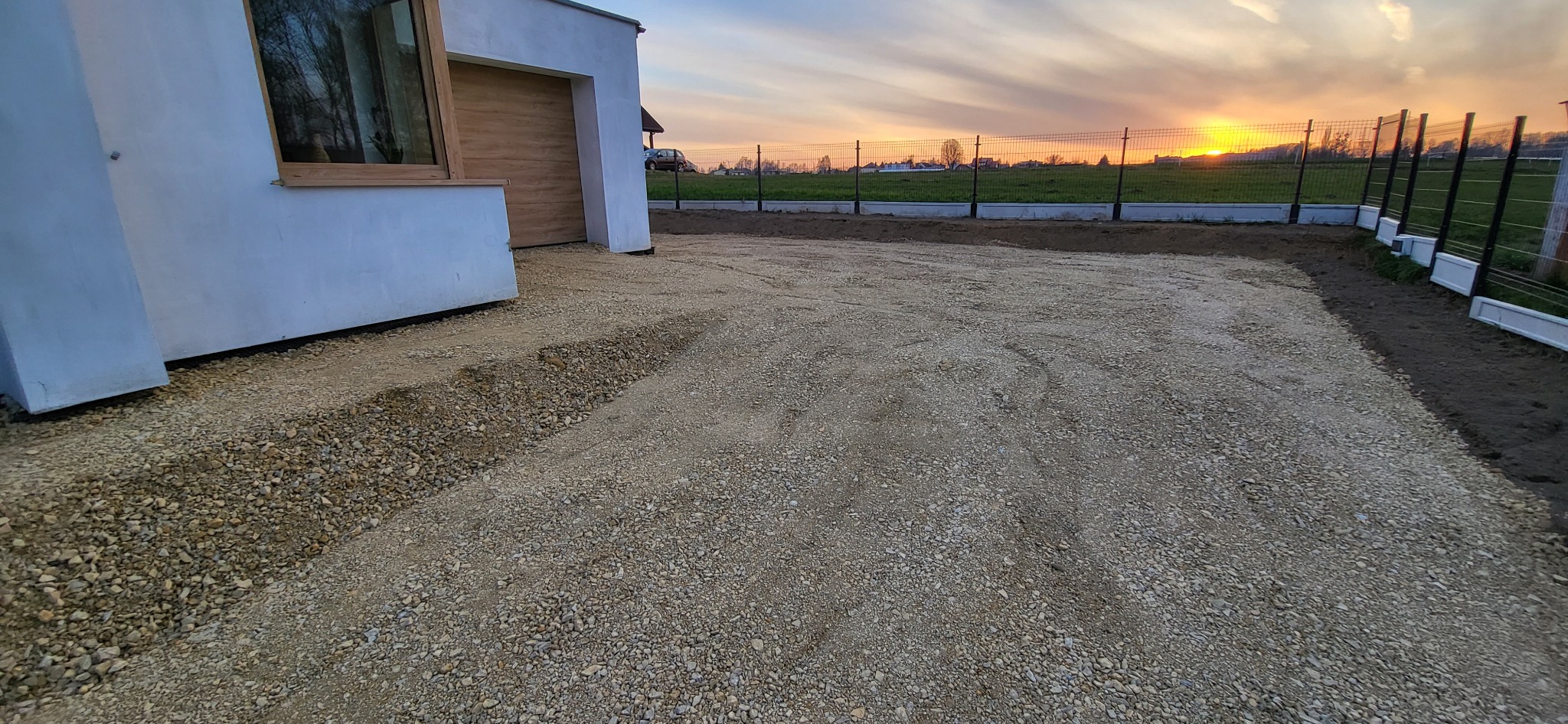Przygotowanie podłoża z kruszywa pod przyszłą nawierzchnię na podjeździe domu jednorodzinnego, widoczne obrzeża trawnika i ogrodzenie panelowe na tle zachodzącego słońca.