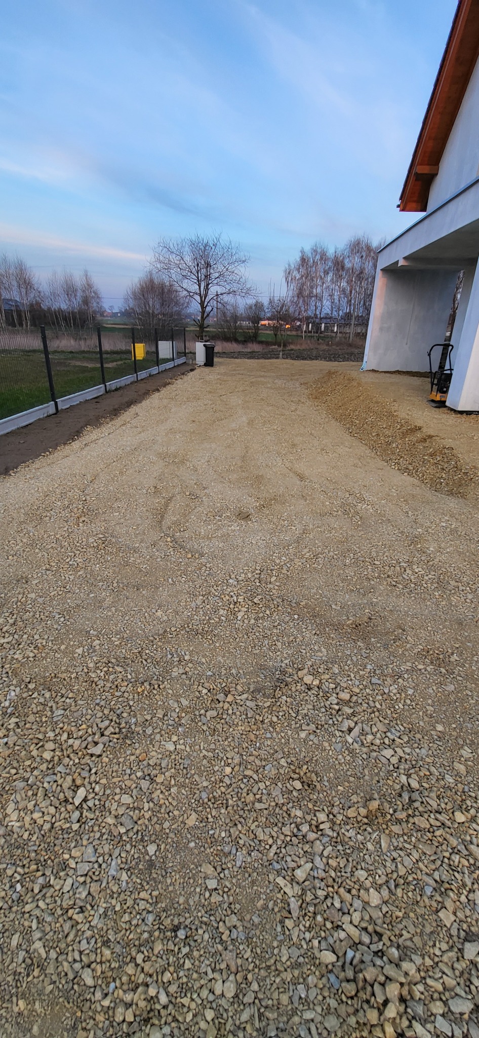 Teren przygotowany pod przyszłą nawierzchnię z kostki brukowej, widoczna warstwa podsypki z kruszywa, obrzeża betonowe i częściowo ogrodzenie panelowe, w tle budynek mieszkalny w stanie surowym...