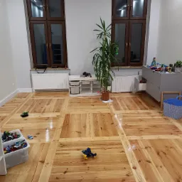 Jasna, drewniana podłoga ułożona w kwadraty z wyraźnym rysunkiem słojów, w pokoju dziecięcym z zabawkami, doniczką z rośliną i meblami w tle.