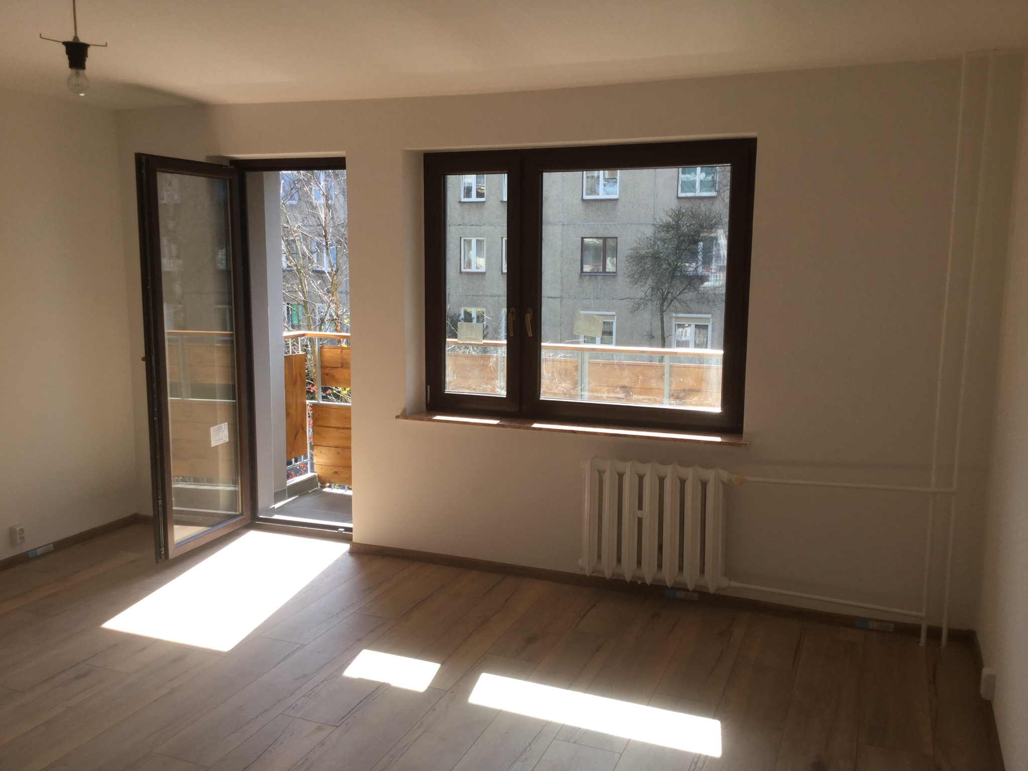 Pusty pokój z drewnianą podłogą, otwartym balkonem i grzejnikiem pod oknem, wpadające promienie słoneczne na podłodze.