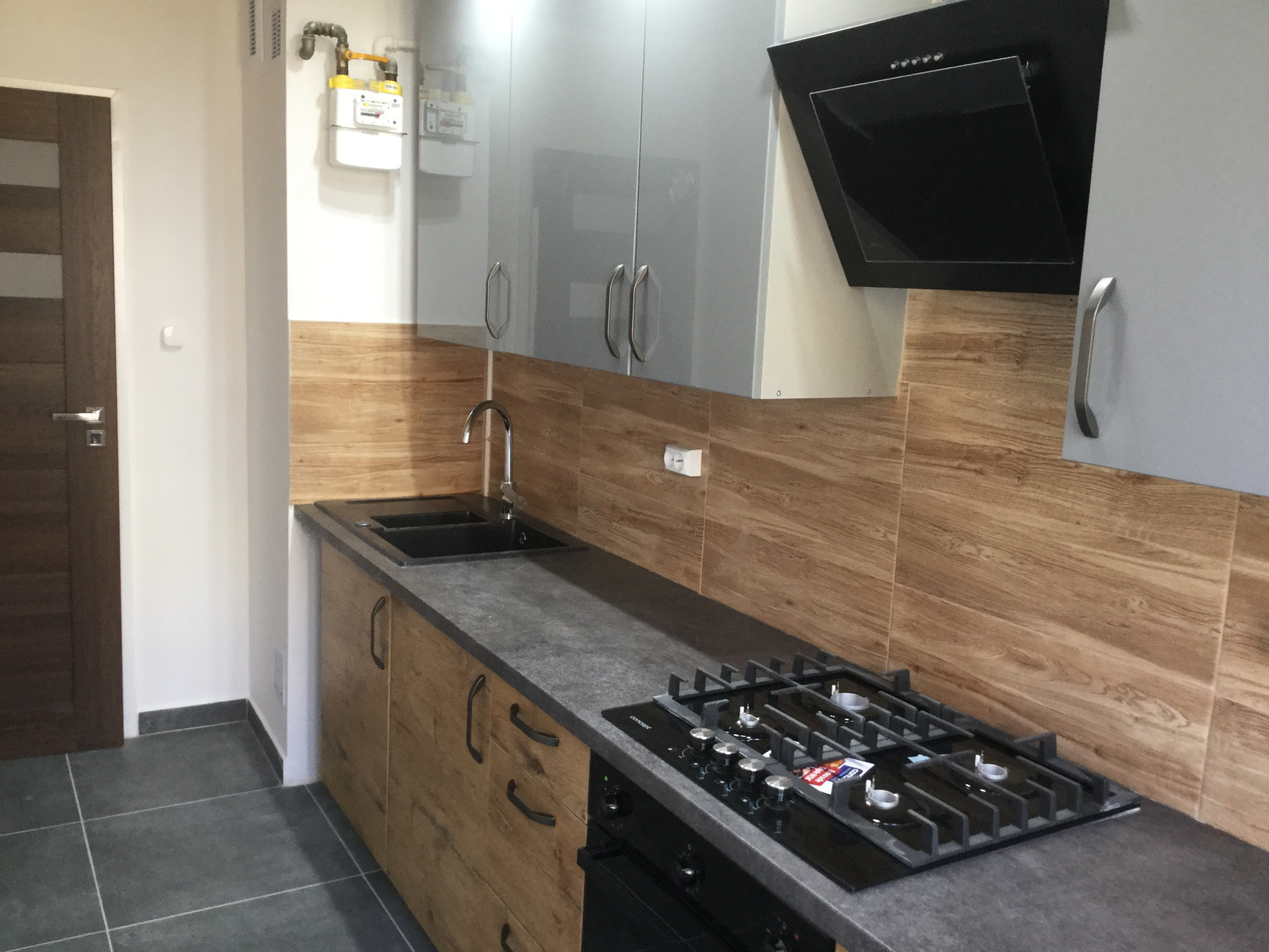 Nowoczesna kuchnia z szarymi szafkami górnymi, drewnopodobnymi dolnymi, czarnym blatem i płytą gazową, drewnopodobne płytki na ścianie, szare płytki podłogowe.