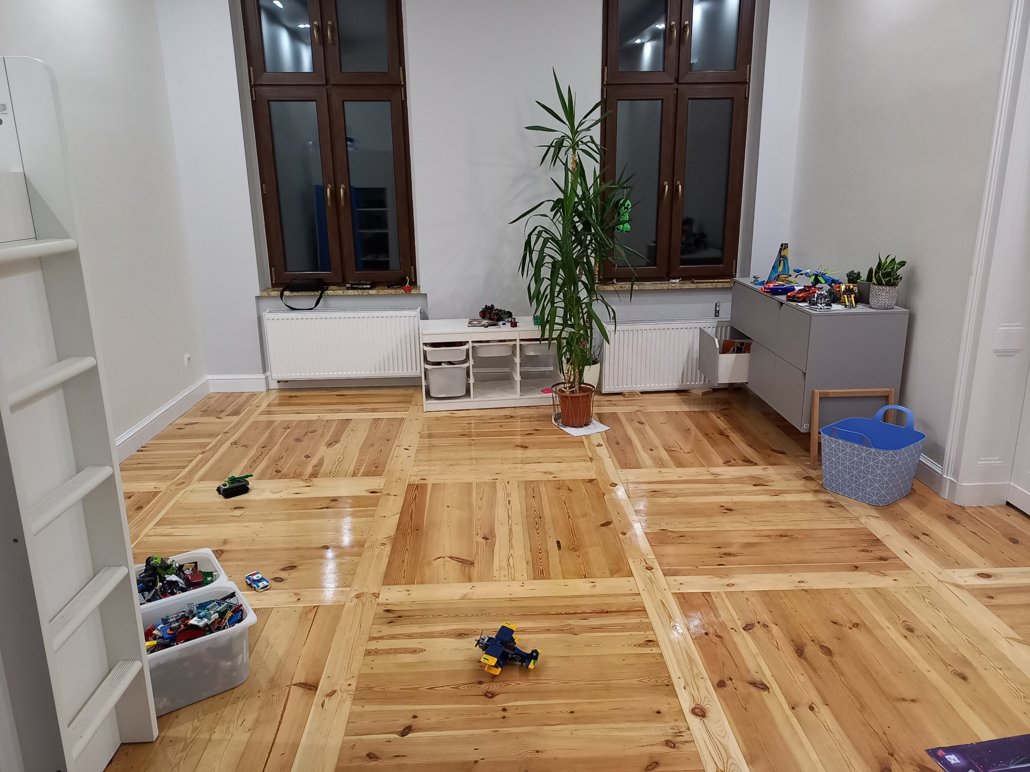 Jasna, drewniana podłoga ułożona w kwadraty z wyraźnym rysunkiem słojów, w pokoju dziecięcym z zabawkami, doniczką z rośliną i meblami w tle.