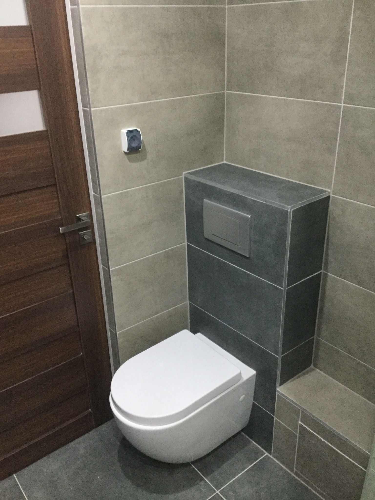 Podwieszana toaleta w nowoczesnej łazience wykończonej szarymi płytkami, z widoczną drewnianą ramą drzwi i minimalistycznym przyciskiem spłukującym.