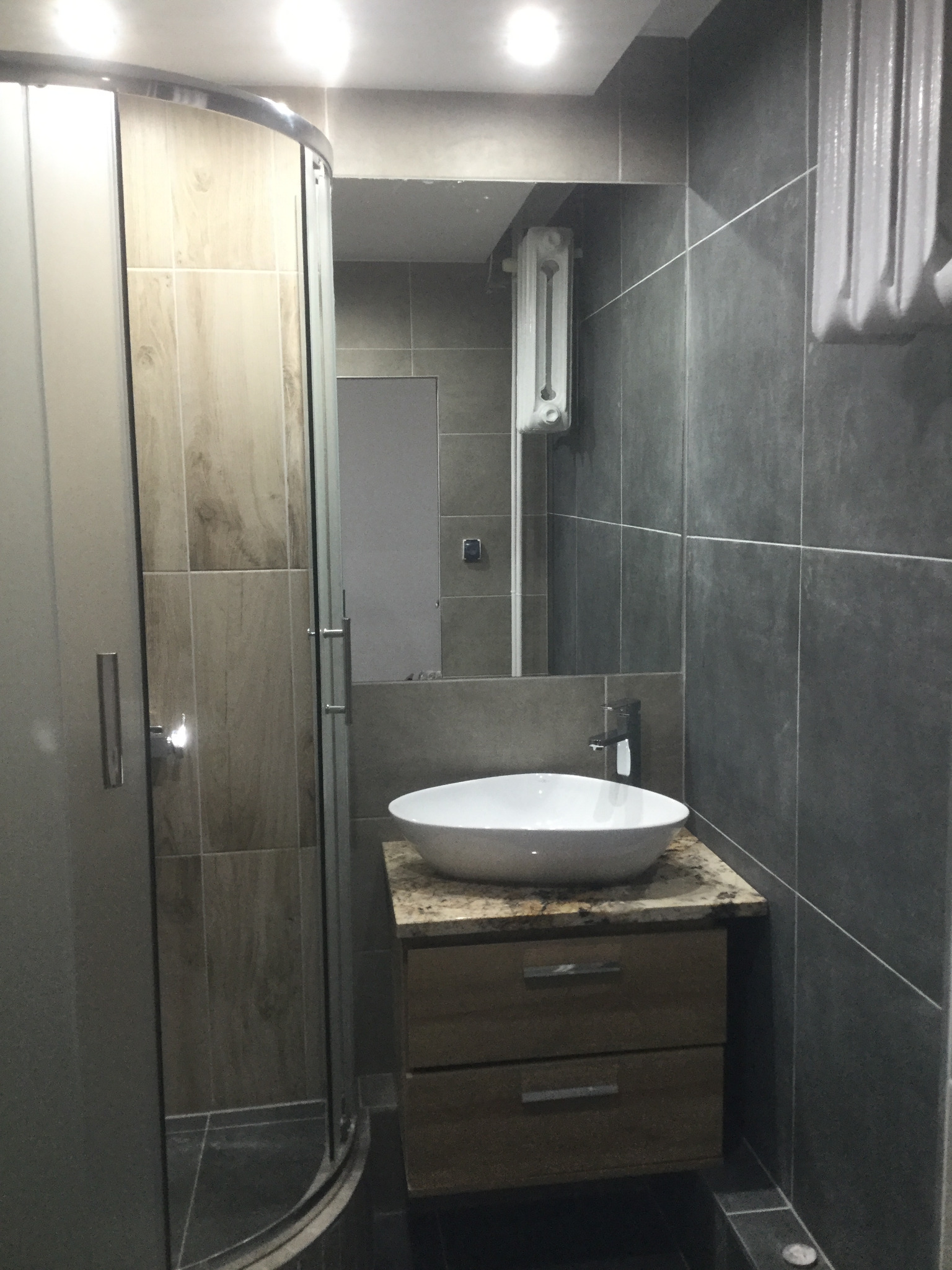 Nowoczesna łazienka z szarą glazurą, kabiną prysznicową z drewnopodobnymi panelami, białą umywalką na szafce z blatem imitującym marmur oraz grzejnikiem na ścianie.