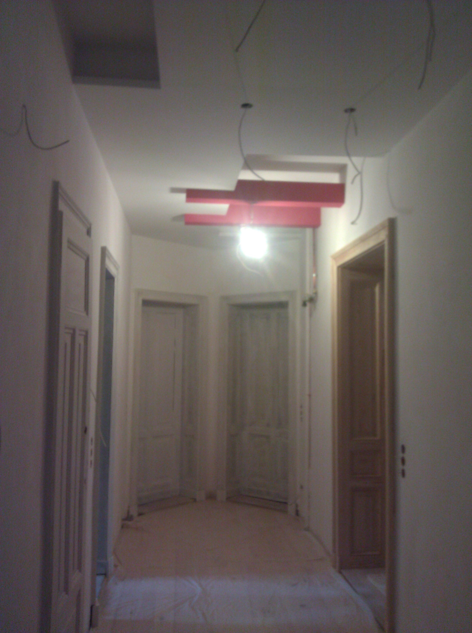 Korytarz w trakcie malowania na biało, z widocznymi czerwonymi elementami dekoracyjnymi na suficie i tymczasowym oświetleniem budowlanym.