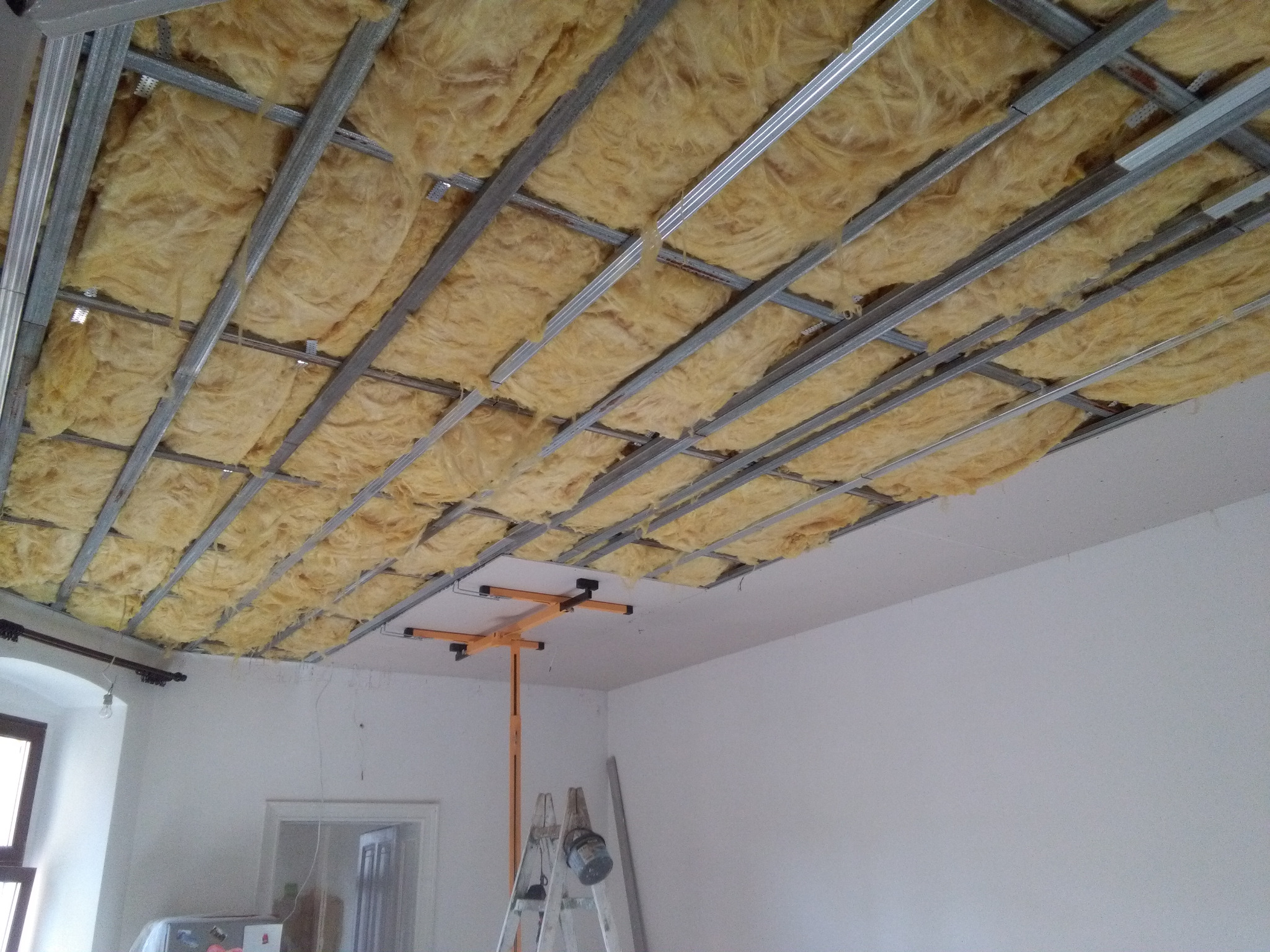 Sufit podwieszany z izolacją z wełny mineralnej, widoczna metalowa konstrukcja nośna i podnośnik do płyt gipsowych, drabina oparta o ścianę w tle.