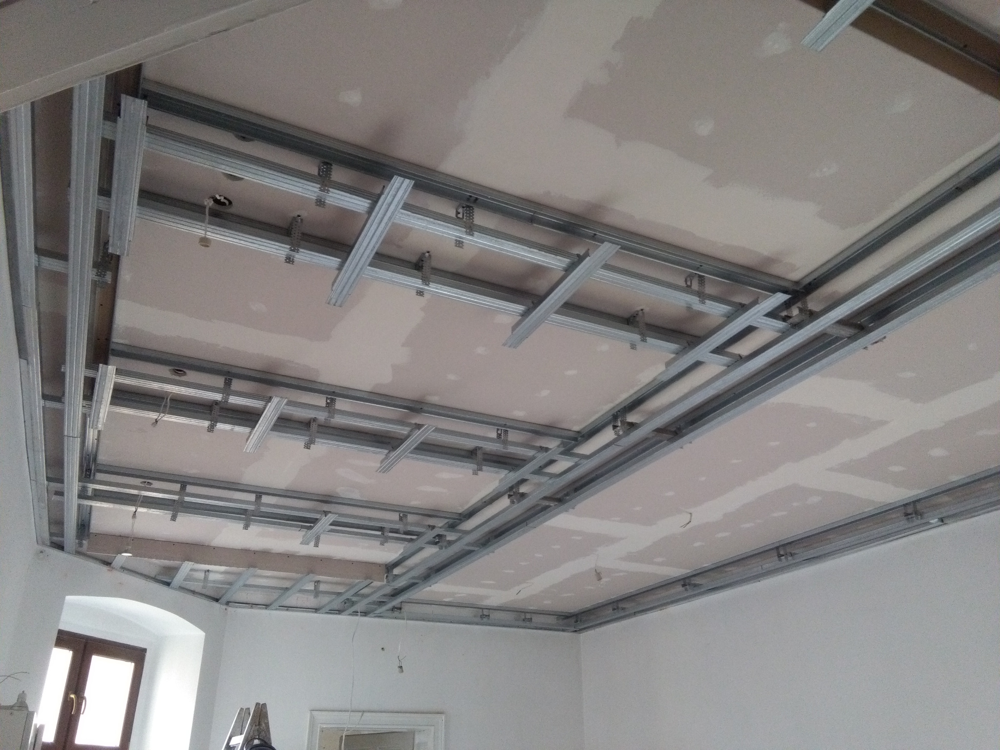 Metalowa konstrukcja sufitu podwieszanego z płyt kartonowo-gipsowych w trakcie montażu, widoczne profile i płyty w trakcie szpachlowania, pomieszczenie z oknem i drabiną w tle.