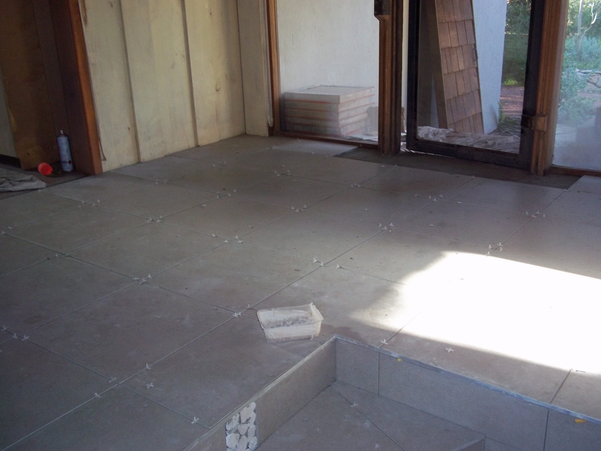 Szare płytki podłogowe ułożone z krzyżykami dystansowymi, widoczny fragment wnęki wyłożonej mozaiką i pudełko z zaprawą, w tle okno z widokiem na zieleń.