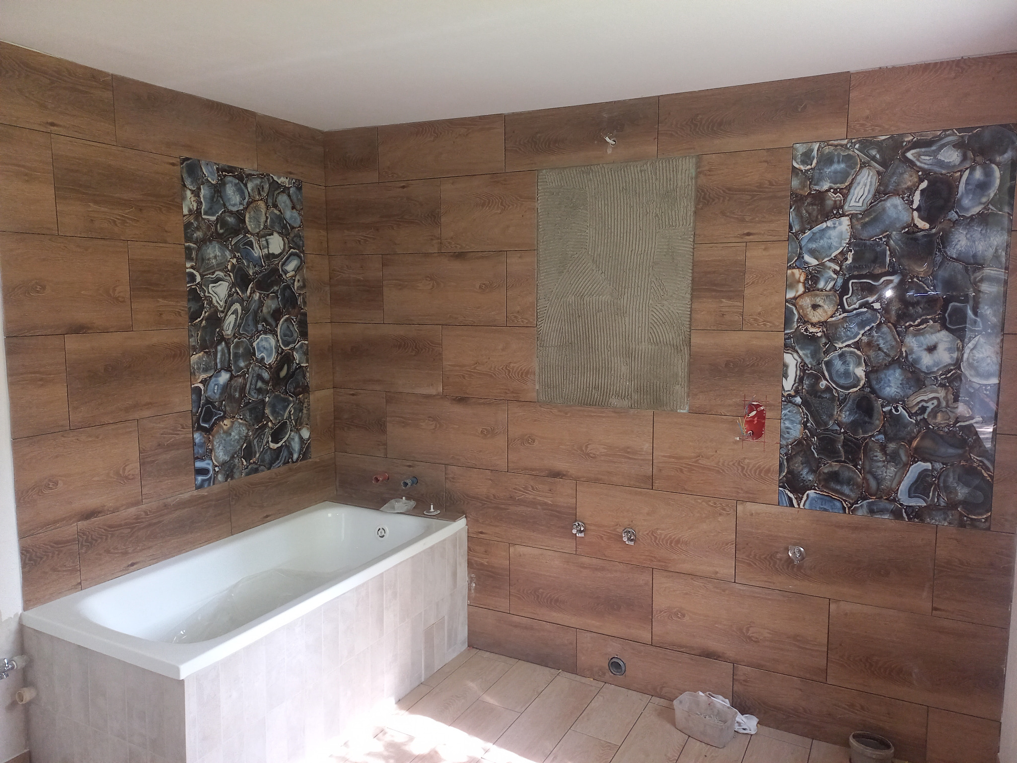 Łazienka w trakcie wykończenia: drewnopodobne płytki ścienne z dekoracyjnymi wstawkami imitującymi agat, biała wanna obudowana jasnymi płytkami, widoczne niedokończone instalacje i przygotowane...