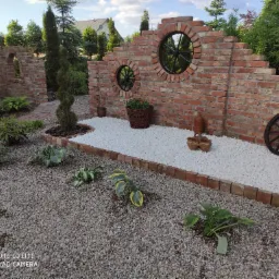 Ogród z ceglanym murem z okrągłymi oknami, białymi kamykami, dekoracjami i roślinami. Ceglana ścieżka w tle.