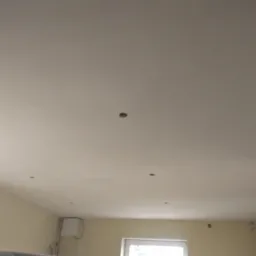 Gładki, jasny sufit po szpachlowaniu i malowaniu, z widocznymi otworami na oświetlenie punktowe, niedoskonałością w prawym górnym rogu i fragmentem ściany w tle.