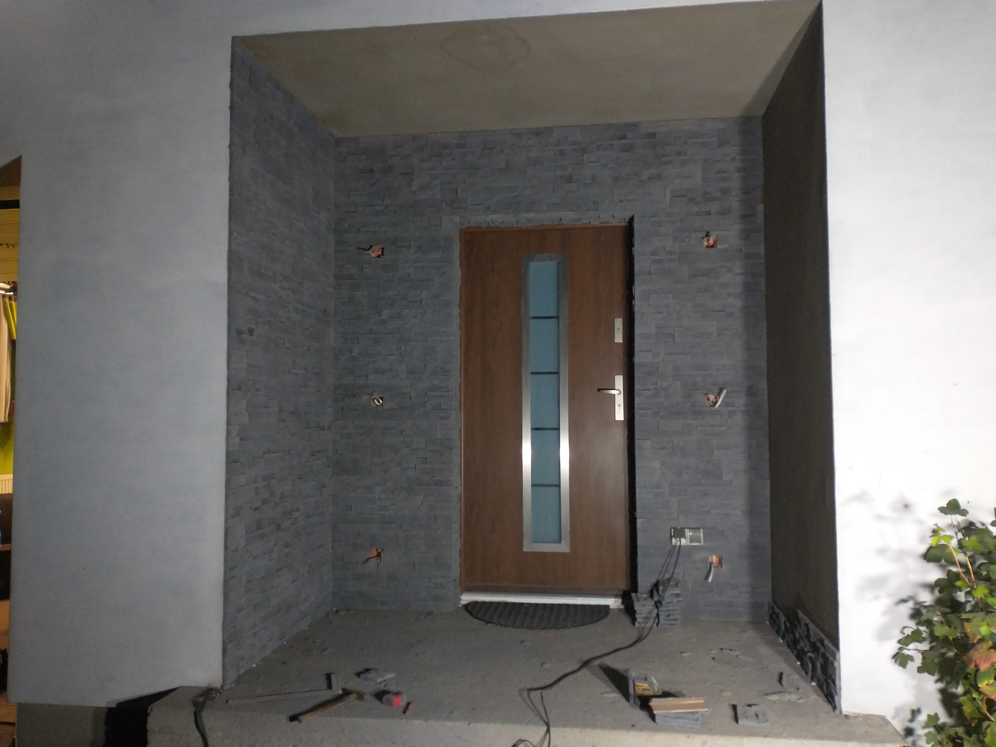 Wejście do budynku w trakcie wykańczania, ściany obłożone szarą kamienną okładziną, widoczne przewody i narzędzia, brązowe drzwi z szybą.