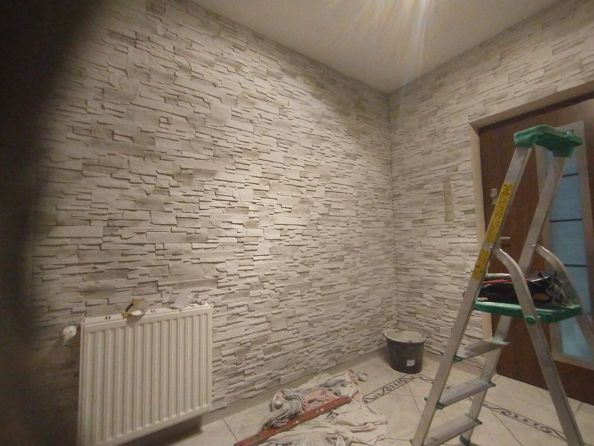 Ściana wykończona dekoracyjną okładziną imitującą kamień, z widocznym grzejnikiem, drabiną i narzędziami malarskimi na podłodze.