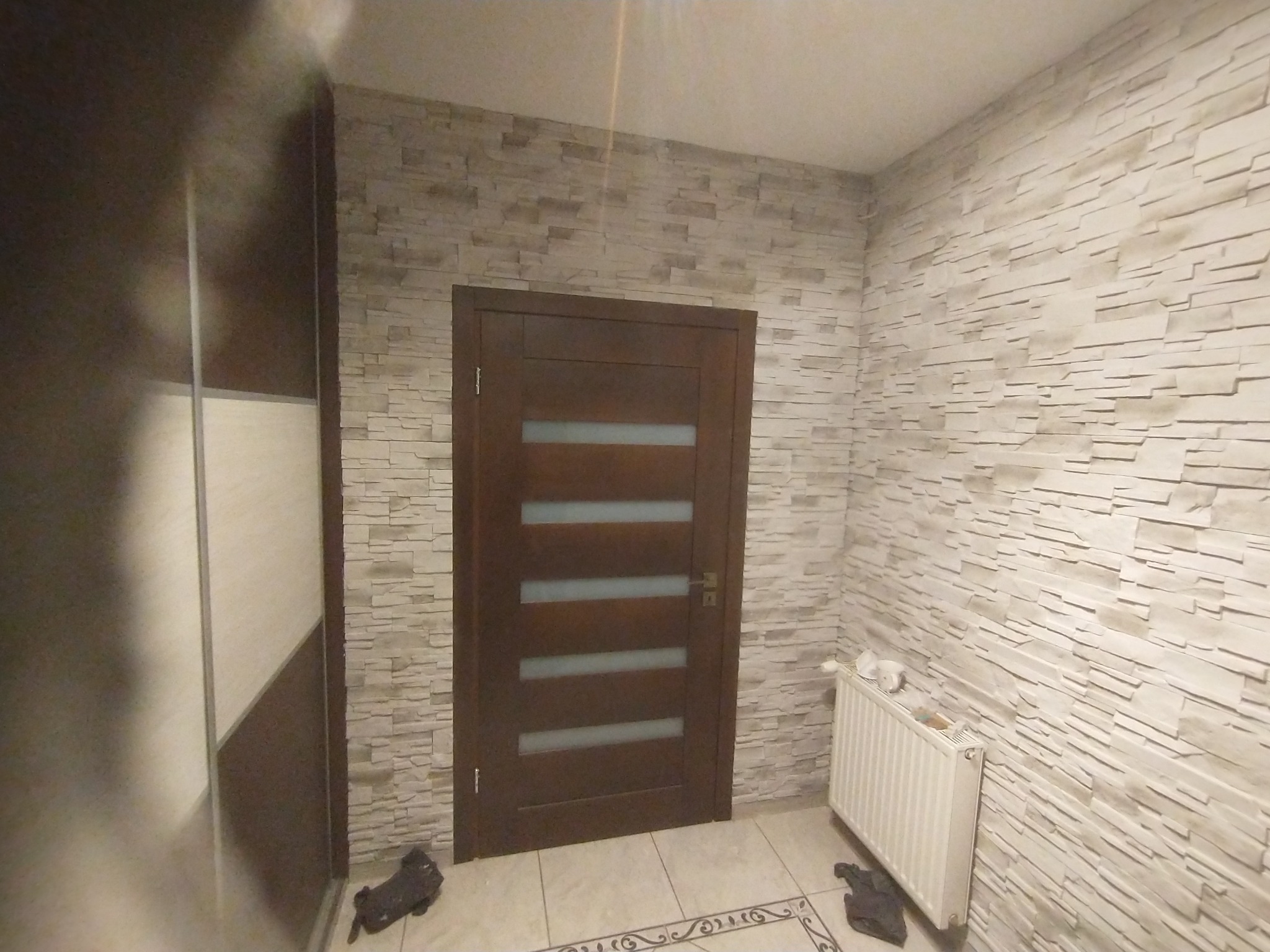Wnętrze przedpokoju z dekoracyjną ścianą z płytek imitujących kamień, brązowe drzwi z przeszkleniami, biały grzejnik i dekoracyjna podłoga z jasnych płytek.
