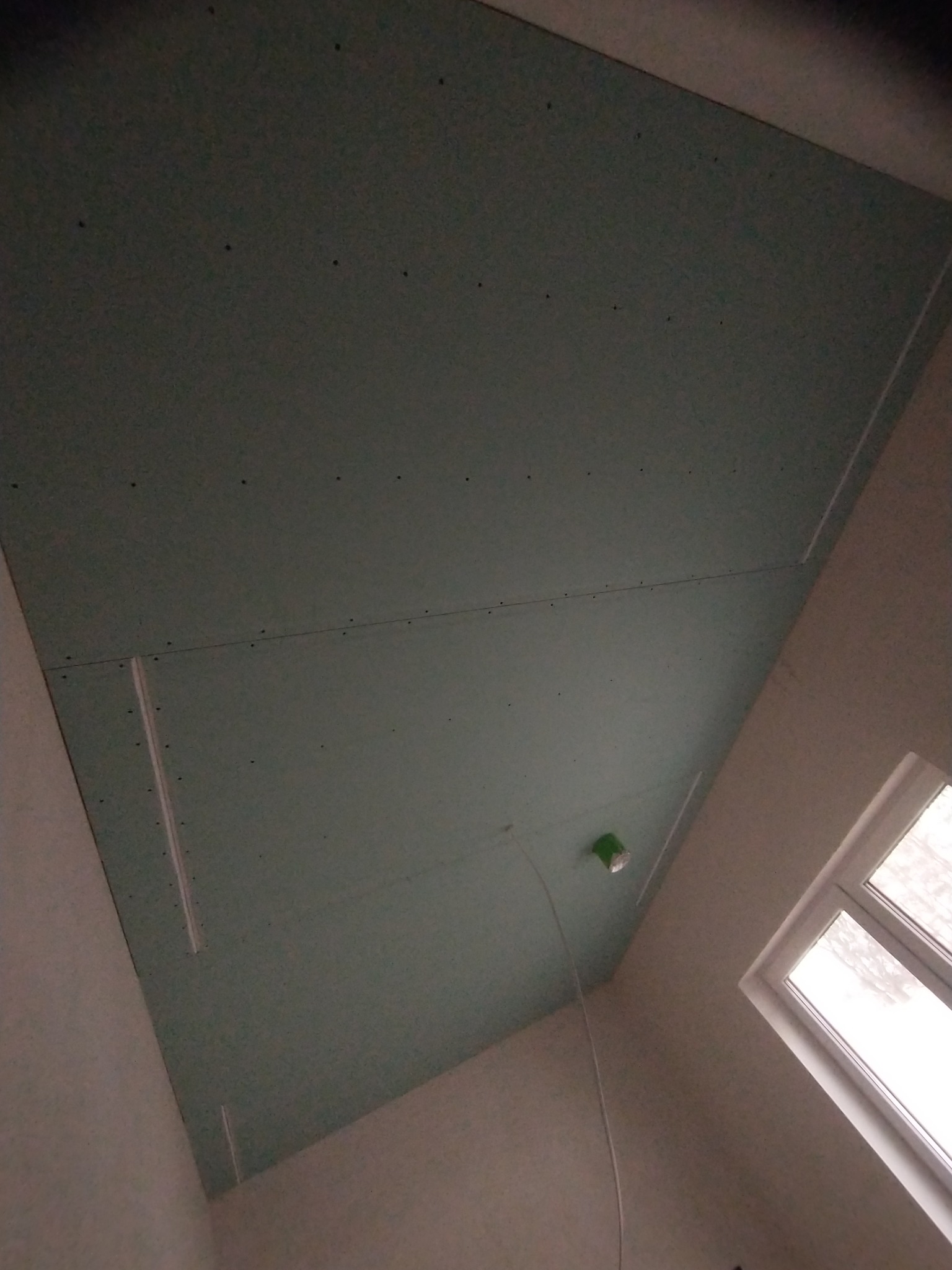 Montaż płyt kartonowo-gipsowych na suficie, widoczne łączenia i śruby mocujące, instalacja elektryczna prowadzona pod płytami, okno w tle.