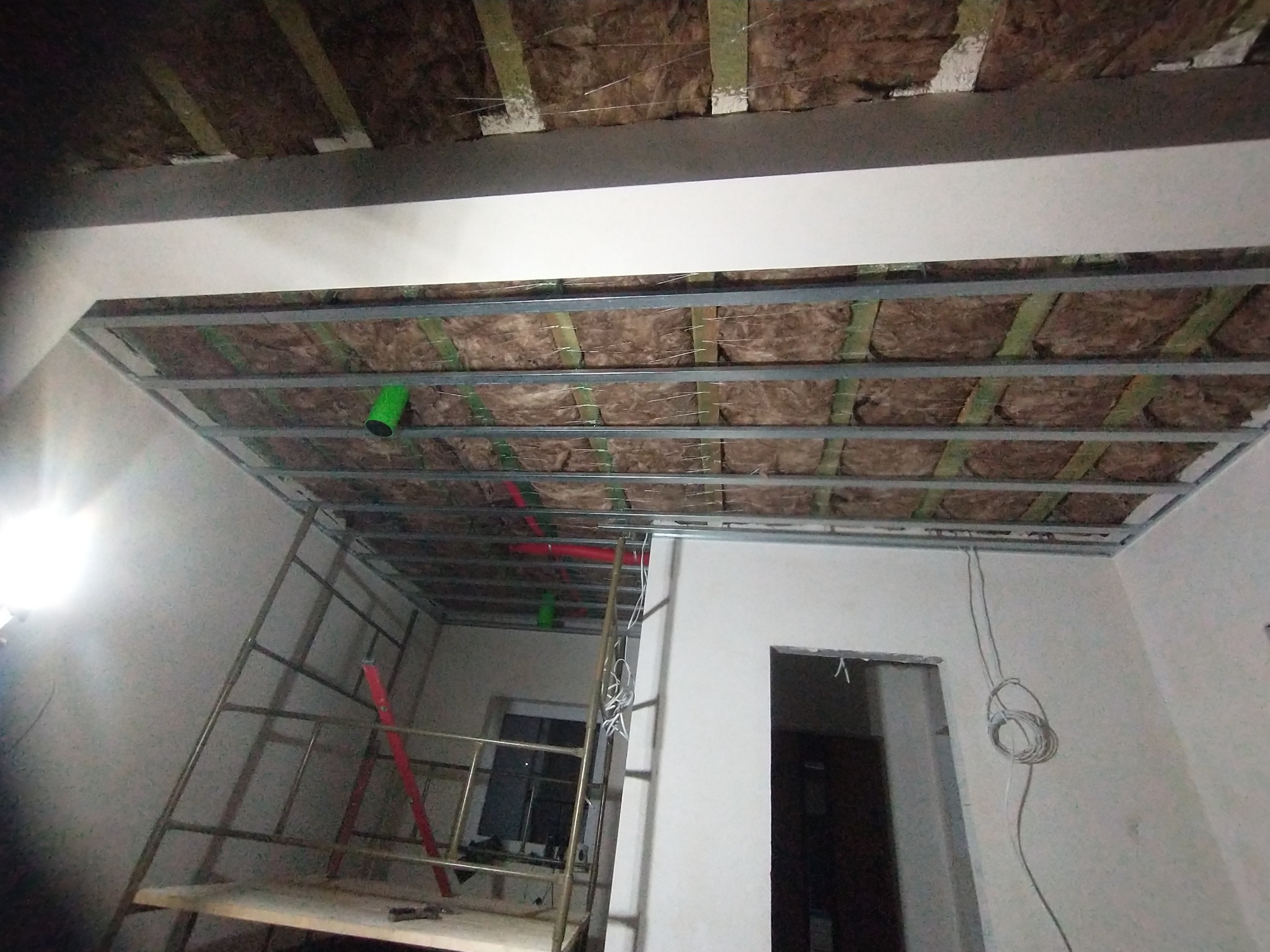 Montaż sufitu podwieszanego z izolacją z wełny mineralnej, widoczna metalowa konstrukcja, rury wentylacyjne i drabina w tle.