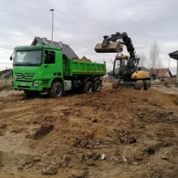 Żółta koparka kołowa nabiera ziemię do łyżki i wysypuje ją do zielonej wywrotki Mercedes na placu budowy z domami w tle.