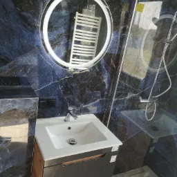 Nowoczesna łazienka z umywalką nablatową, okrągłym lustrem z podświetleniem LED i kabiną prysznicową, wykończona płytkami imitującymi ciemny marmur.