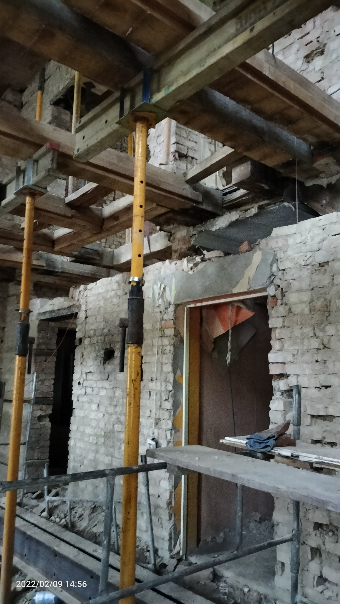 Wnętrze budynku w trakcie rozbiórki, widoczne ceglane ściany, drewniane belki stropowe podparte żółtymi stemplami budowlanymi, fragment rusztowania i otwór drzwiowy.