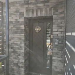 Wejście do domu z elewacją z szarej cegły klinkierowej, ciemne drzwi z szybą, dekoracja w kształcie serca na drzwiach, torba z bułkami na klamce, widoczne puszki elektryczne na elewacji.