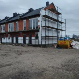 Budynek szeregowy w trakcie prac elewacyjnych: część budynku ocieplona styropianem i otoczona rusztowaniem, część z odsłoniętą czerwoną cegłą, widoczny kontener ze śmieciami budowlanymi.