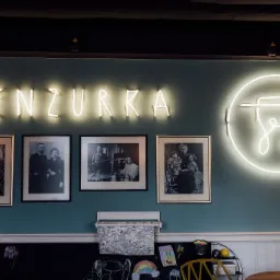 Turkusowa ściana z neonowym napisem MENZURKA i okrągłym neonem z logo, podświetlona lampą z żarówką w kształcie kolby, udekorowana czarno-białymi fotografiami w ramkach i rysunkami kredą na czarnej...